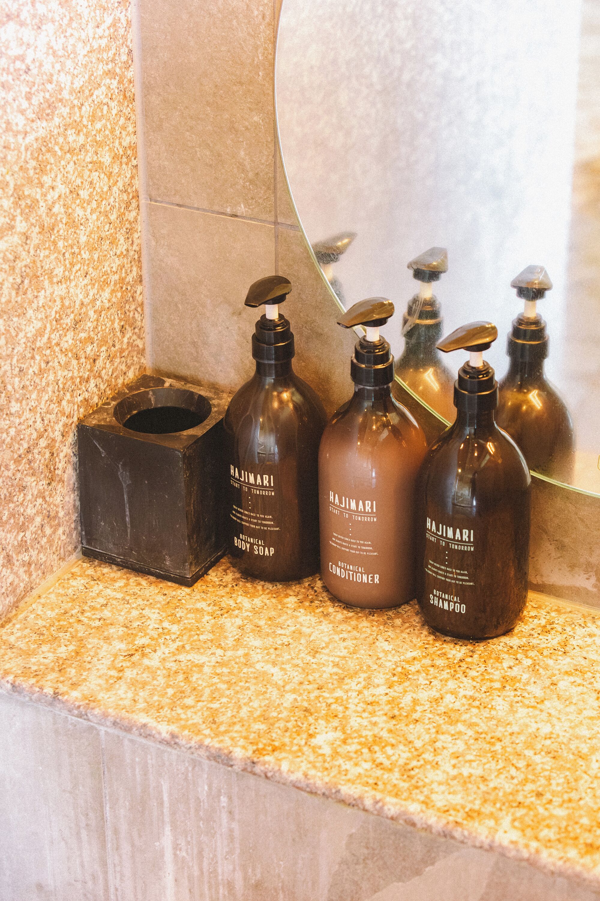 Un meuble-vasque de salle de bain avec trois flacons de savon pour le corps de la marque Hajimari et un porte-savon noir.