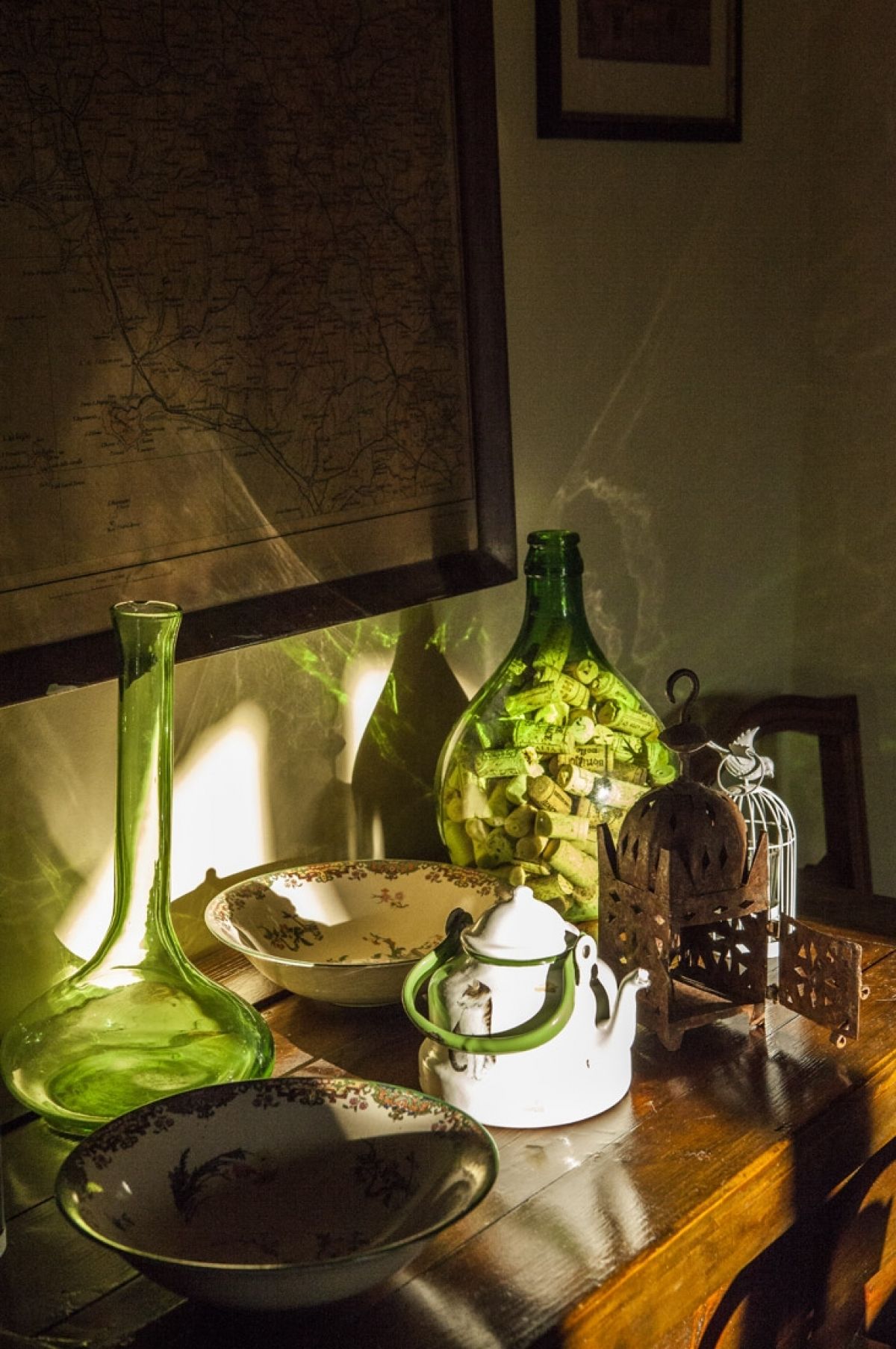 Una mesa con un mapa, una botella de vidrio verde, una tetera blanca y una jaula de pájaros de metal.