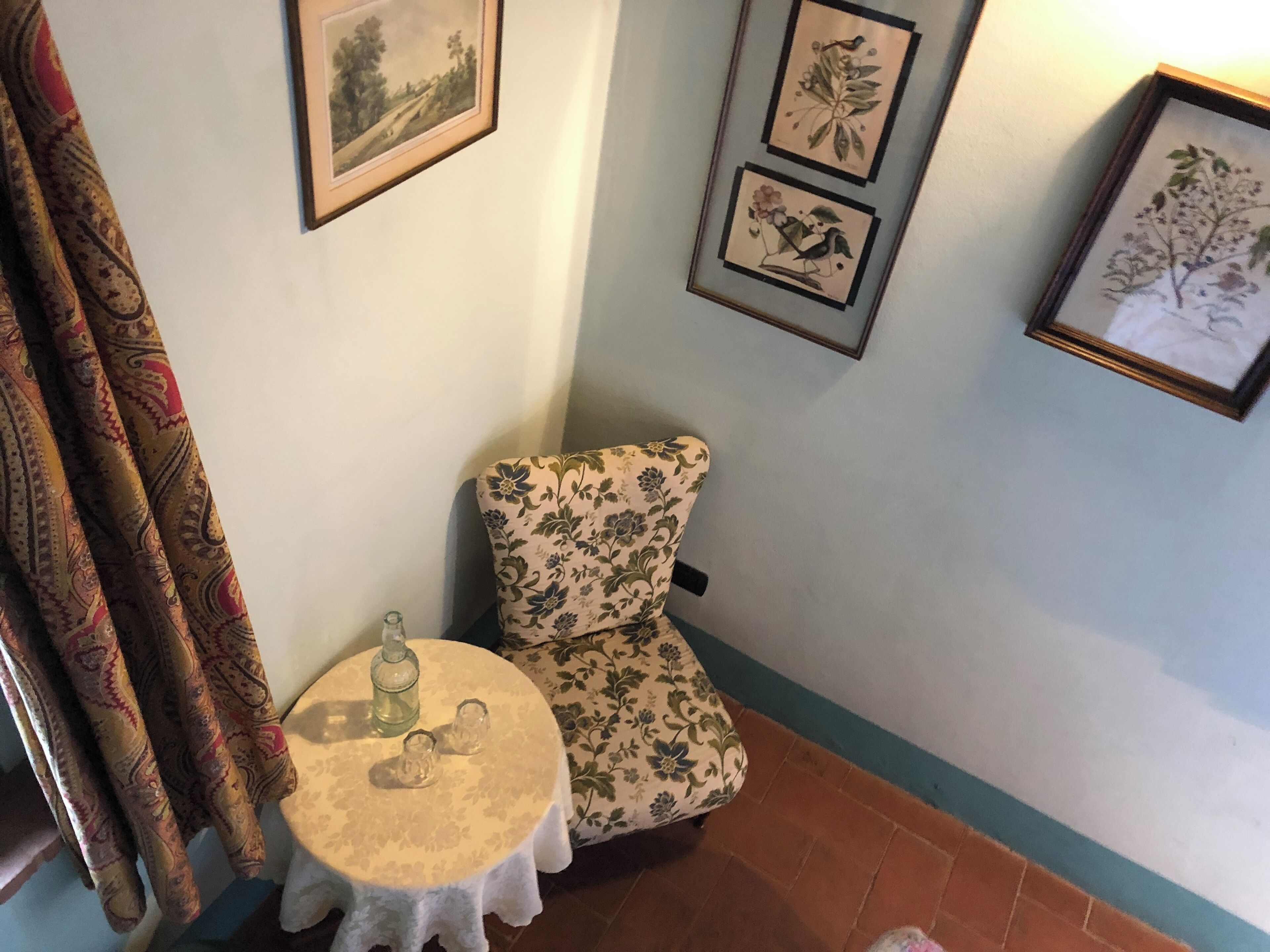 Una habitación de esquina con un sillón estampado con motivos florales, una mesita redonda con una botella y vasos, y tres láminas botánicas enmarcadas en la pared.