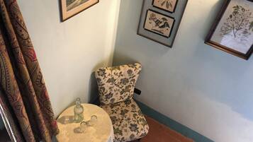 Una habitación de esquina con un sillón estampado con motivos florales, una mesita redonda con una botella y vasos, y tres láminas botánicas enmarcadas en la pared.
