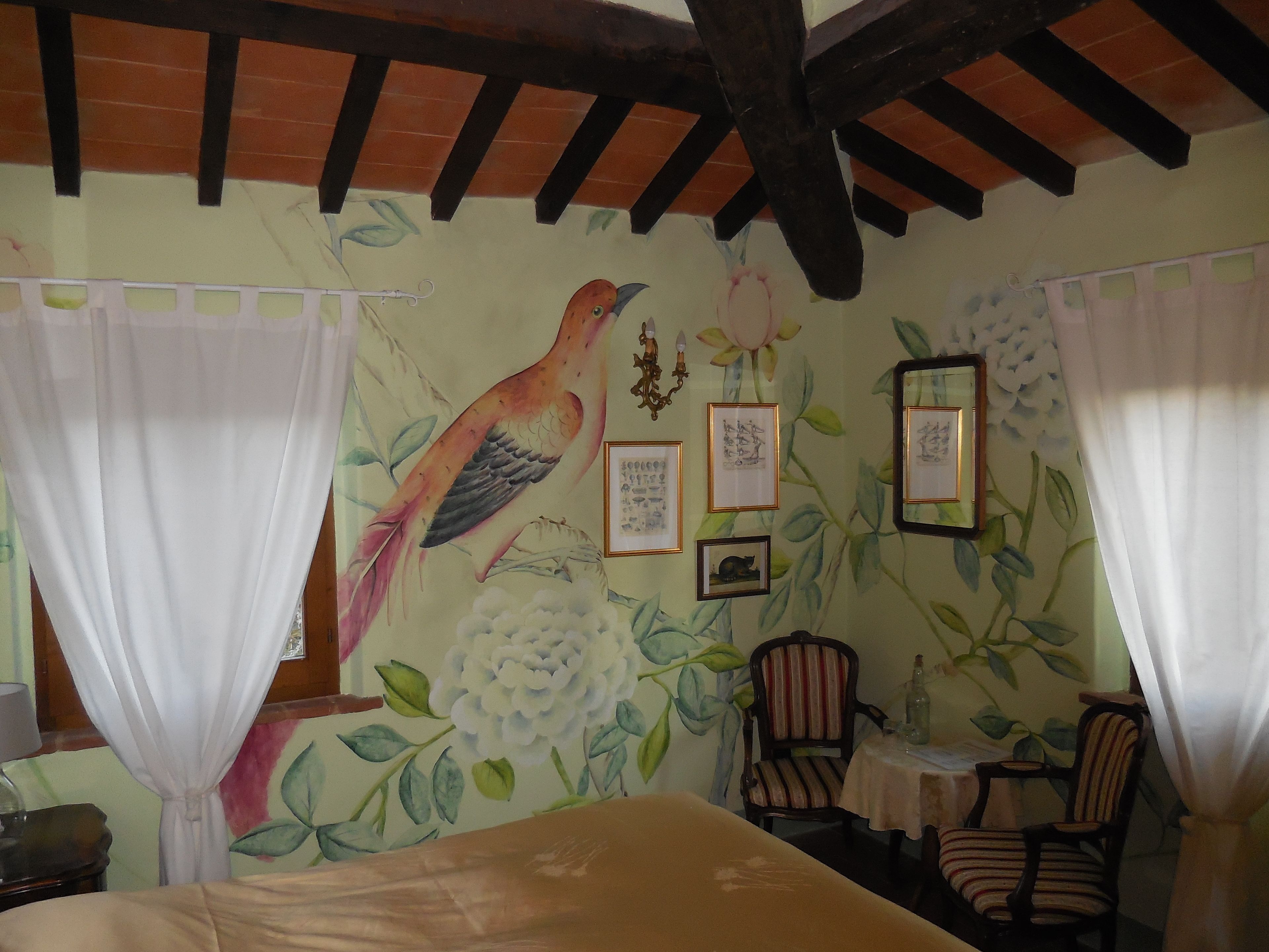 Una habitación con cama, una silla, una mesa y un muro decorado con motivos florales y de pájaros.