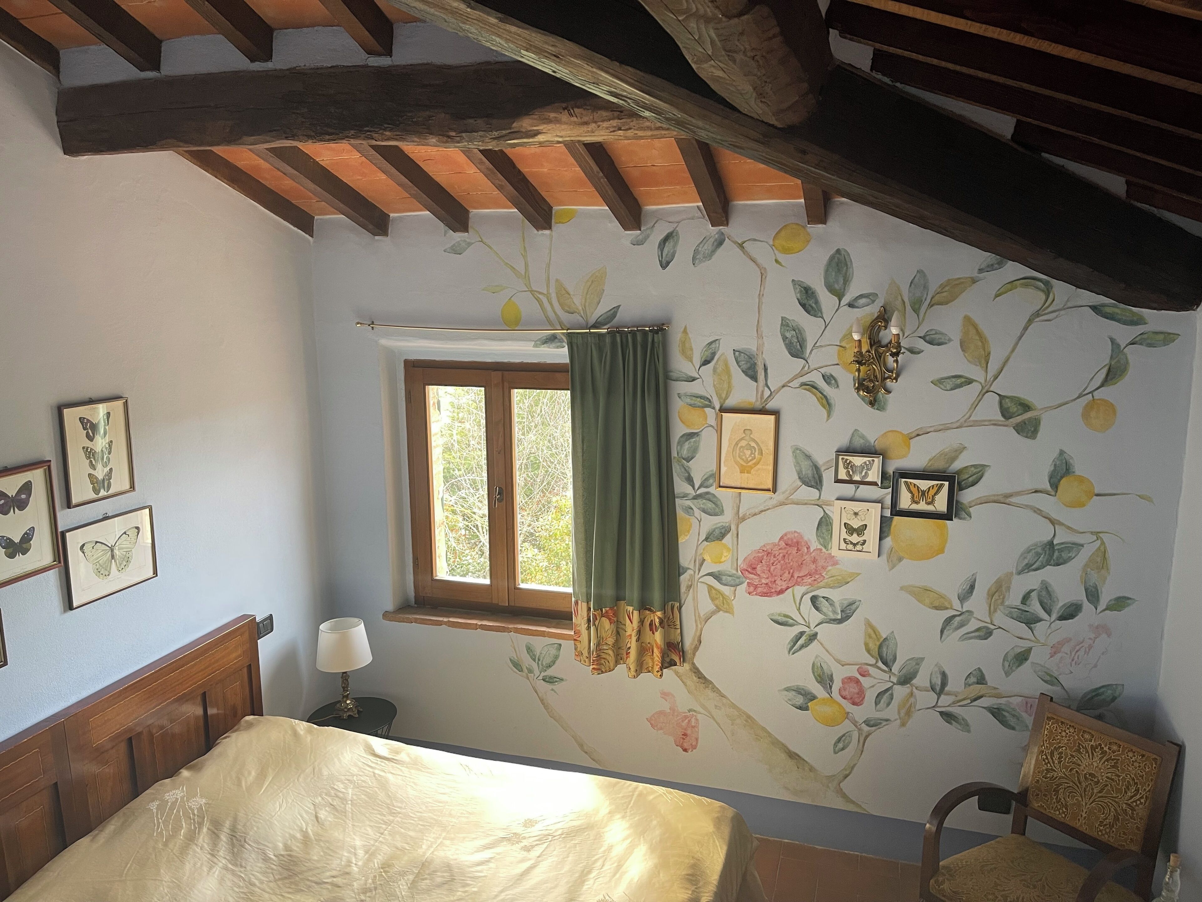 A bedroom with a bed, a window with a curtain, and a wall with framed pictures and a floral mural.