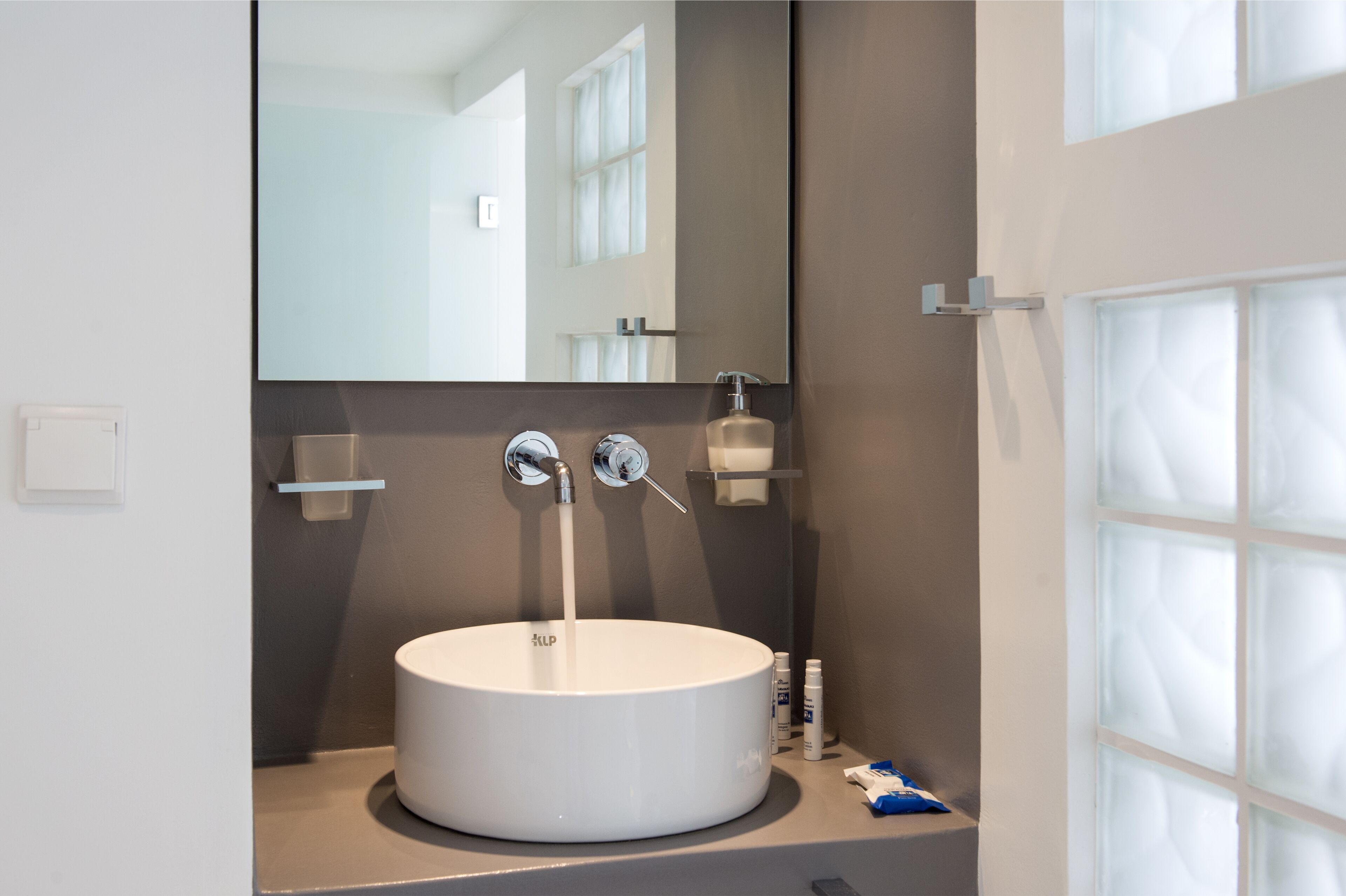 A modern bathroom with a round white sink, a mirror, and a window with frosted glass panels.