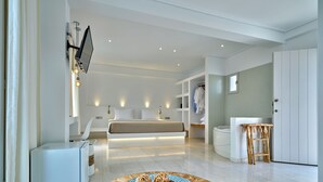 A modern bedroom with a large bed, bedside tables, a round coffee table with decorative items, a wardrobe, and a bathroom with a bathtub and towel rack. - Kanale's Rooms and Suites (Paros)