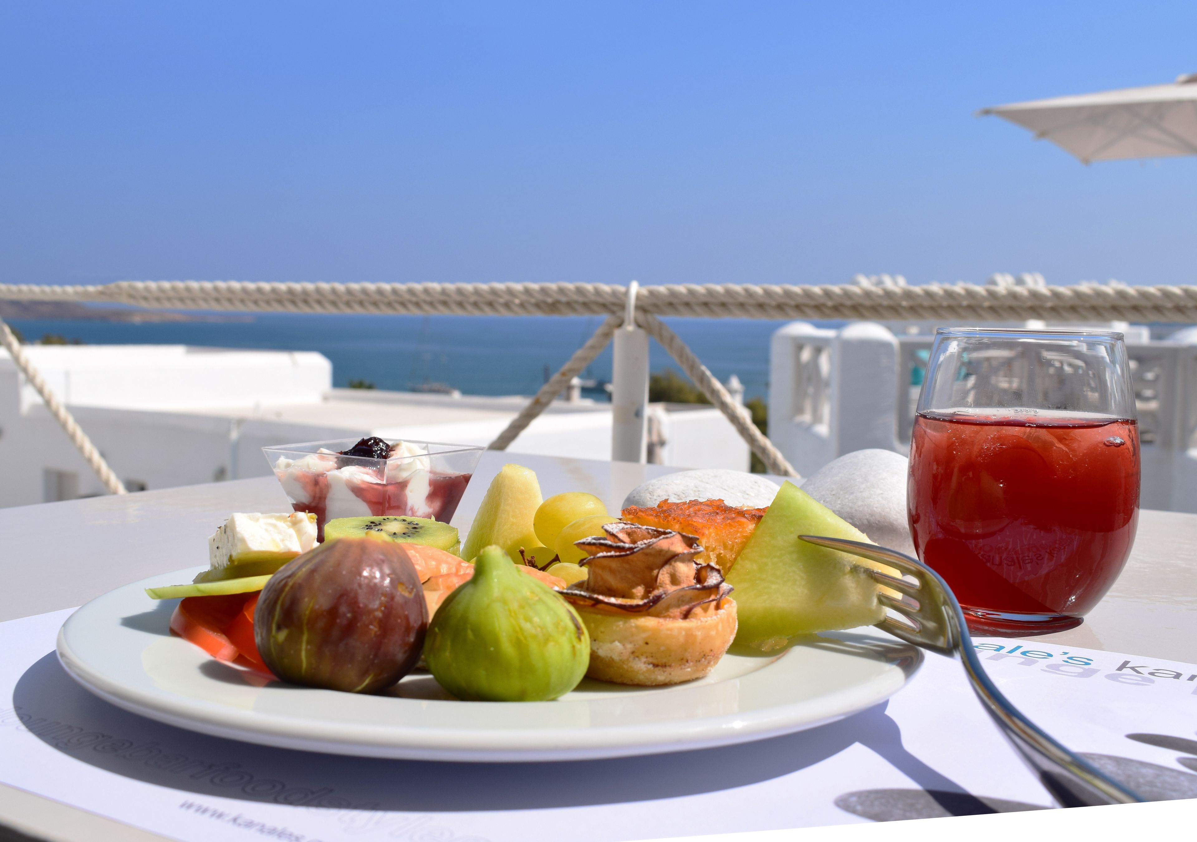 A table with a plate of assorted fruits and a glass of red drink, set against a clear blue sky and ocean view.