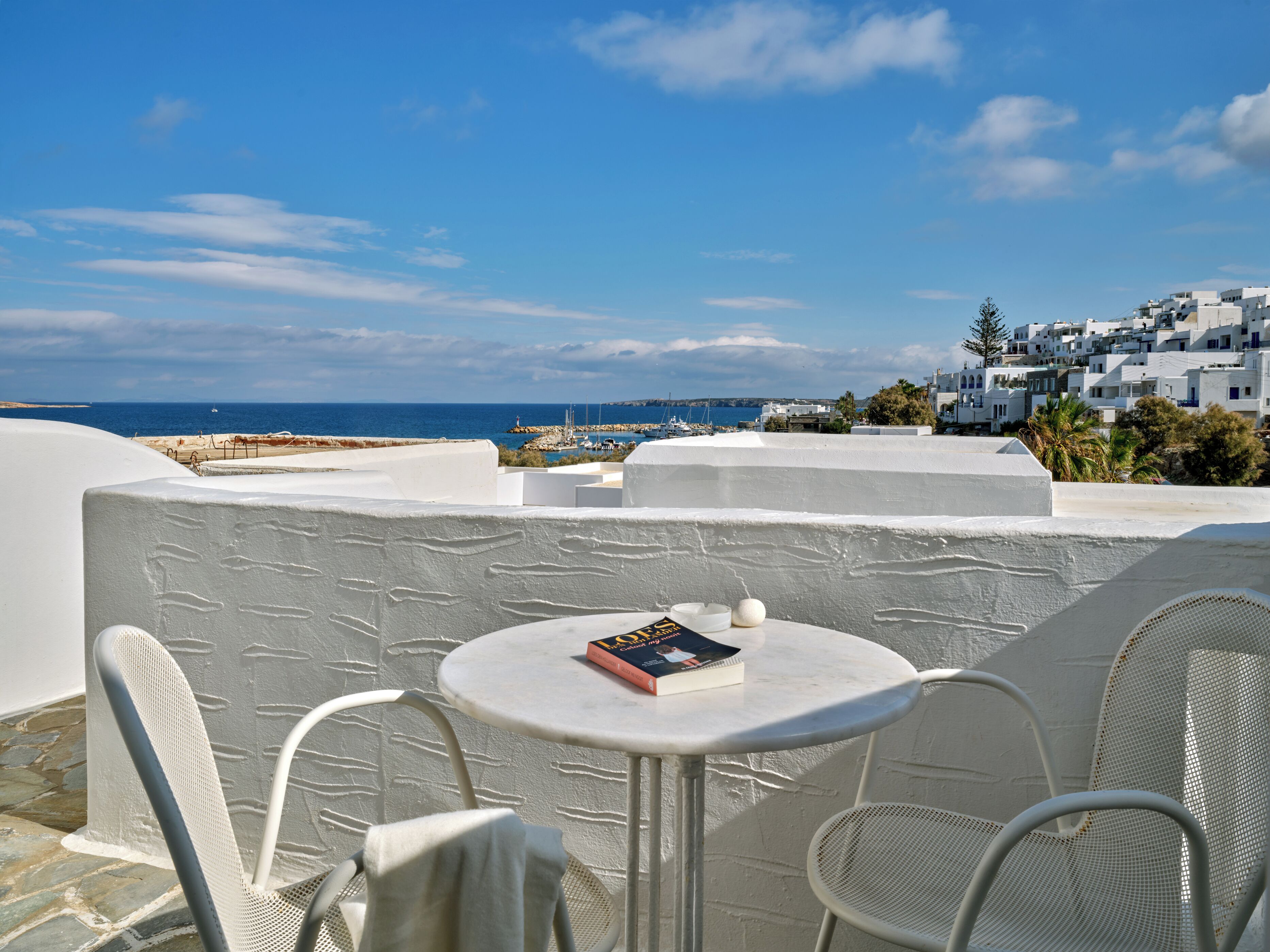 A white-walled terrace with a round table and two chairs, overlooking a coastal town and the sea.