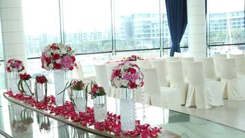 Zona de recepción con una mesa de vidrio decorada con arreglos florales y pétalos de rosa, sillas blancas y ventanas amplias con vista a la ciudad.