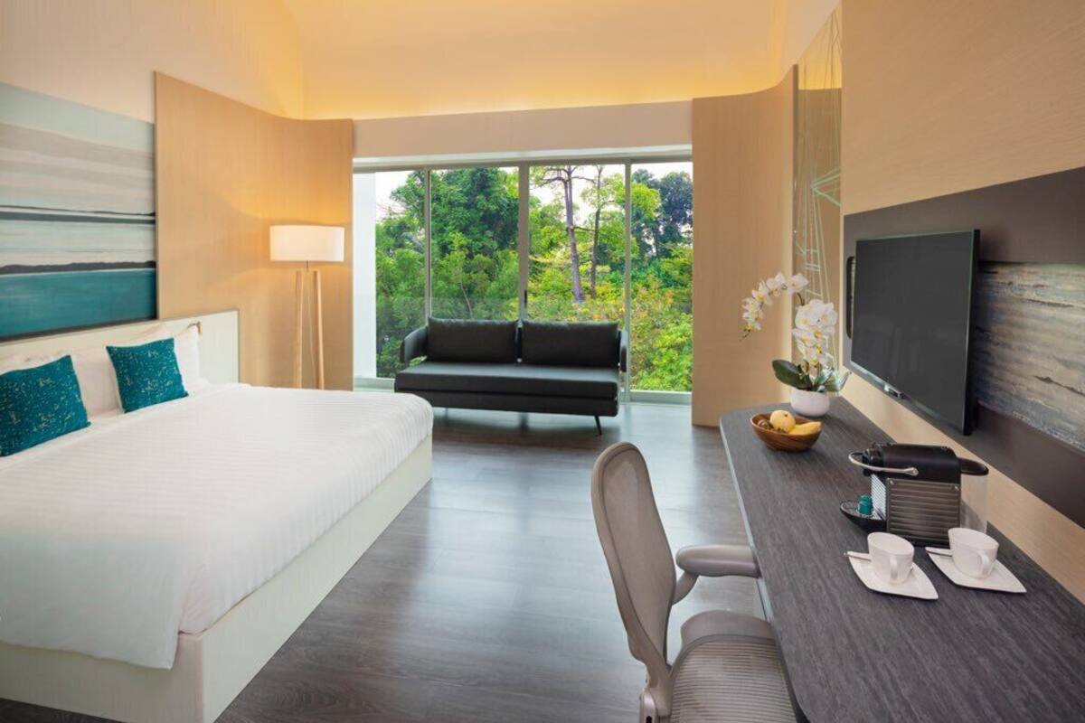 Une chambre d’hôtel moderne dotée d’un grand lit, d’un canapé, d’un bureau avec une chaise, d’une télévision et offrant une vue sur la verdure environnante.