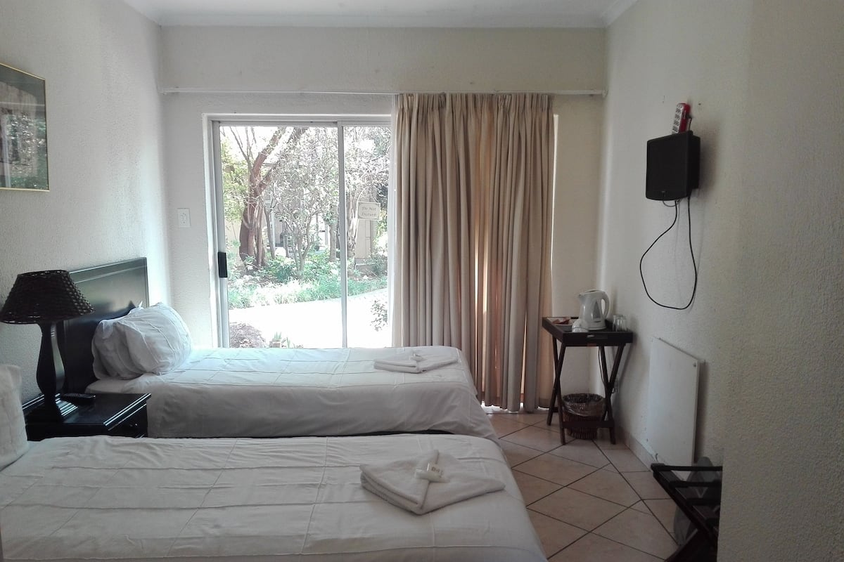 A hotel room with two beds, a bedside table with a lamp, a small table with a kettle, and a TV mounted on the wall.