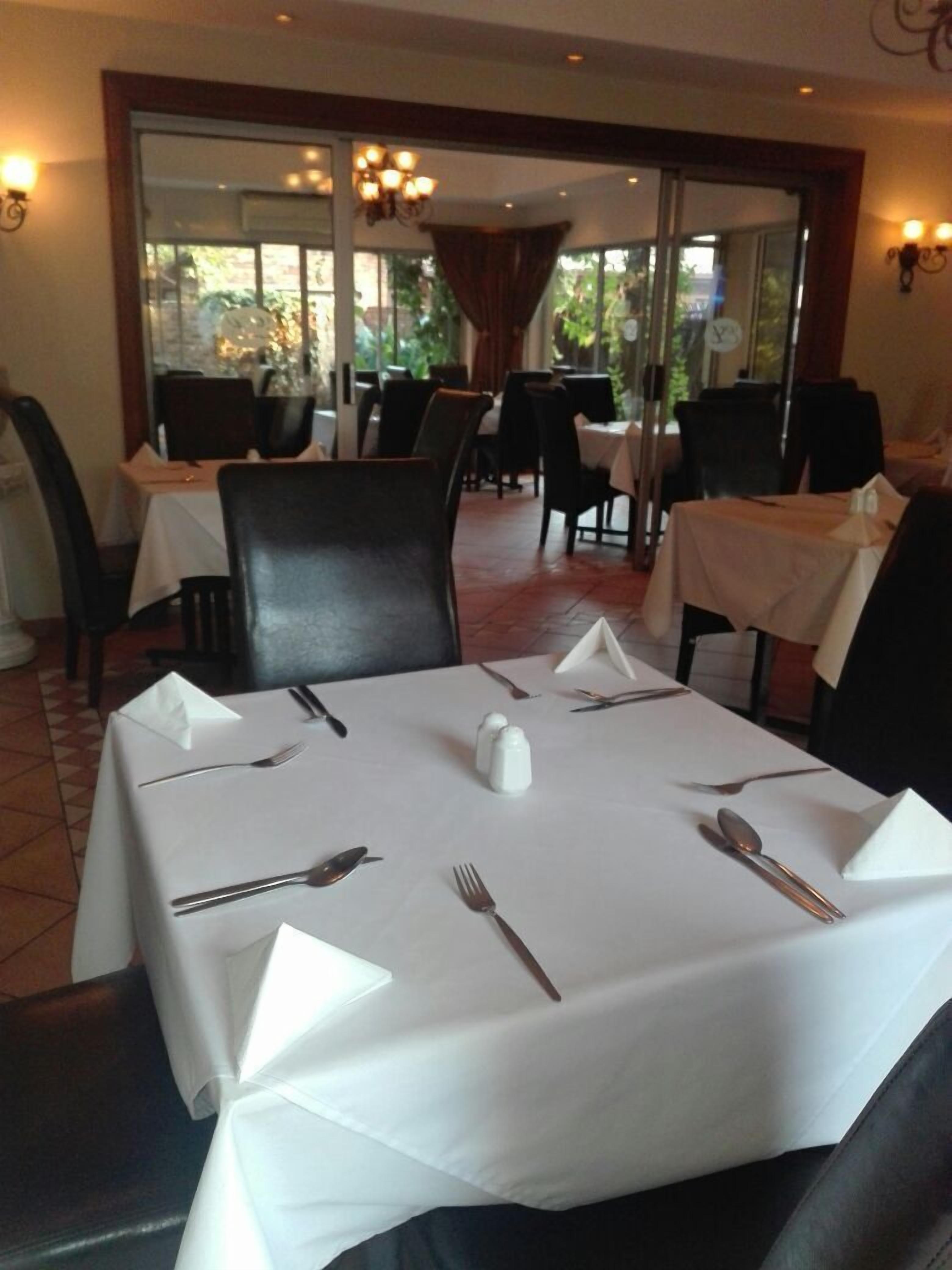 A dining area with tables set for four, each with white tablecloths, silverware, and salt and pepper shakers. Chairs are arranged around the tables.