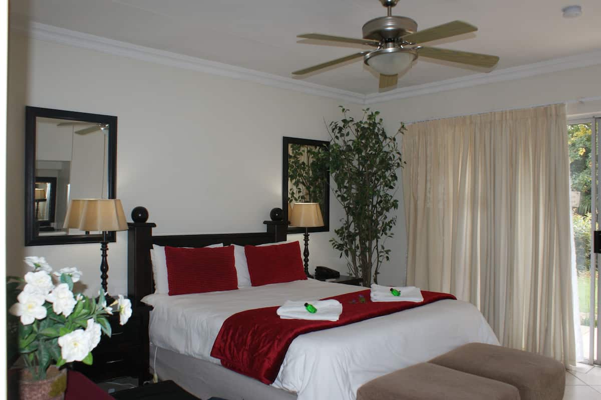 A hotel room with a bed, a ceiling fan, a large mirror, a plant, and a window with curtains.