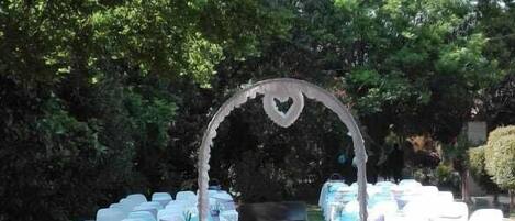 Une arche décorative pour cérémonie de mariage installée en plein air, avec un tapis rouge, entourée de chaises et de tables blanches, le tout aménagé dans un jardin arboré.