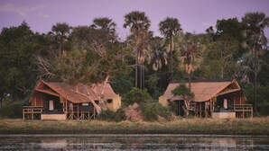 Dos cabañas rústicas con techos de paja, rodeadas de densa vegetación tropical y una masa de agua tranquila.