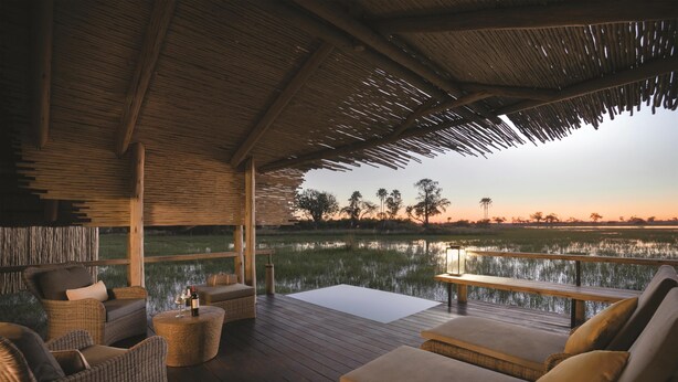 Un área de descanso al aire libre con mobiliario de mimbre, una terraza de madera y vista a un cuerpo de agua al atardecer.