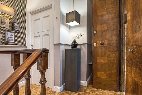 Una escalera de madera con barandilla, una puerta blanca con el número 5 arriba, un jarrón negro con flores blancas sobre un pedestal, y una puerta de madera con manija metálica.