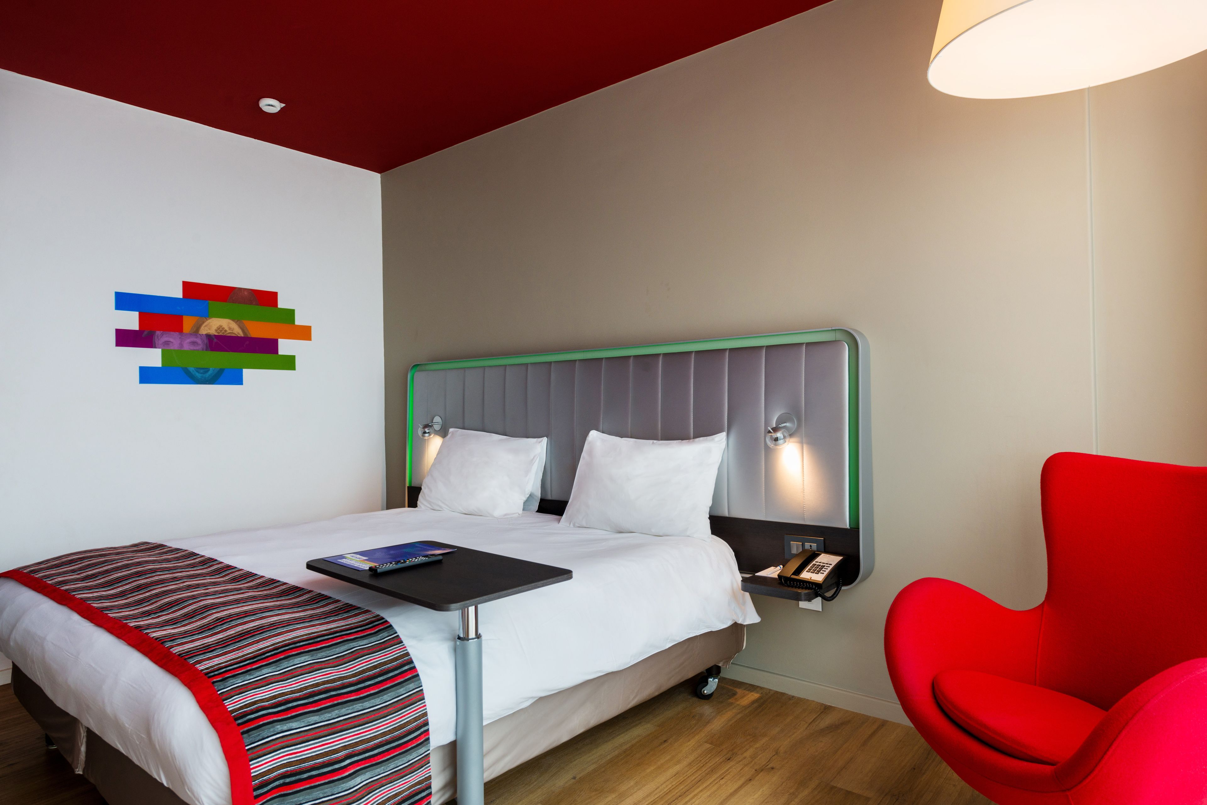 Habitació d'hotel amb un llit, una cadira vermella, una taula de nit amb un telèfon i una obra d'art a la paret.