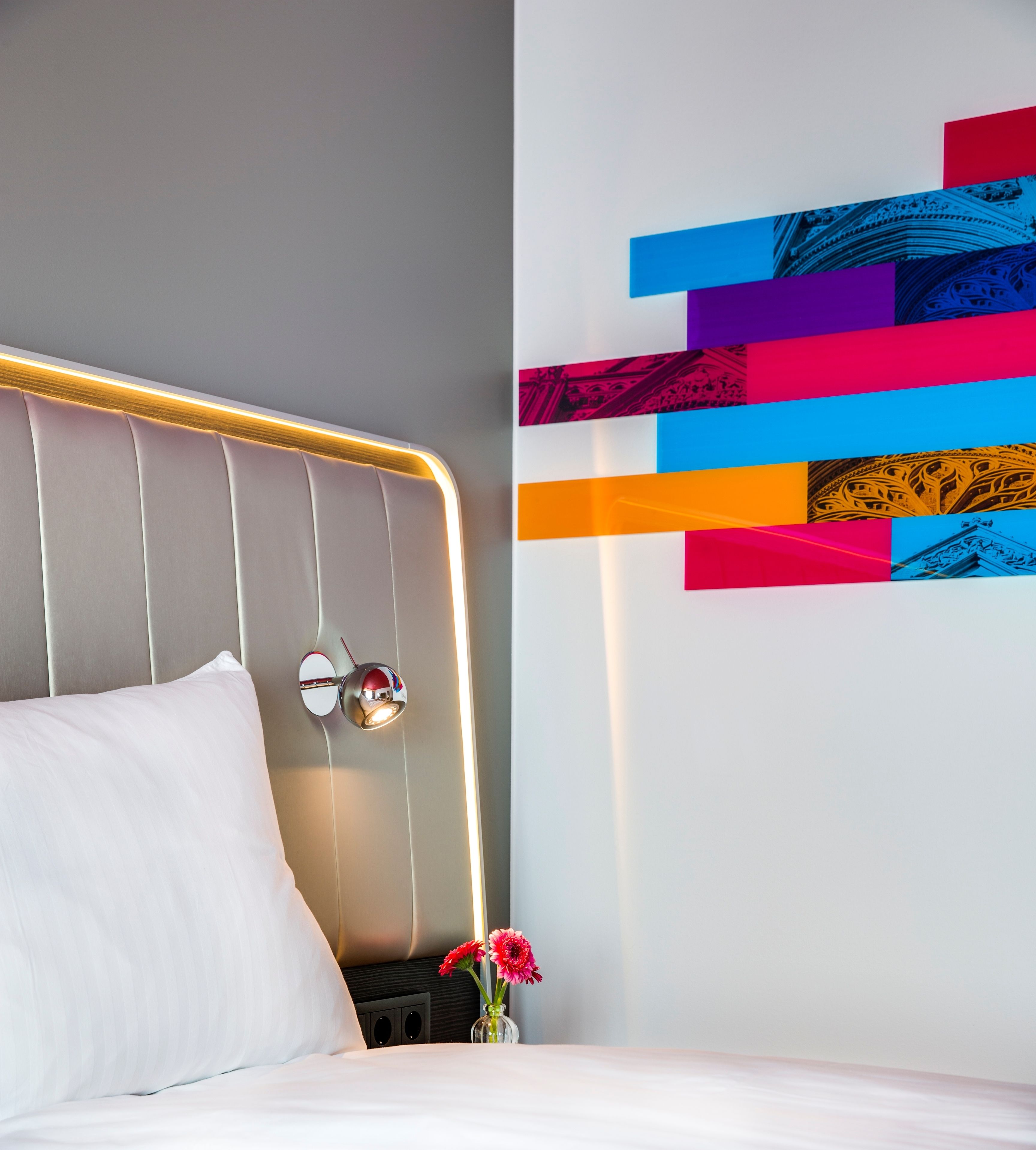 A modern hotel room with a neatly made bed, a wall-mounted light fixture, and a colorful abstract art piece.