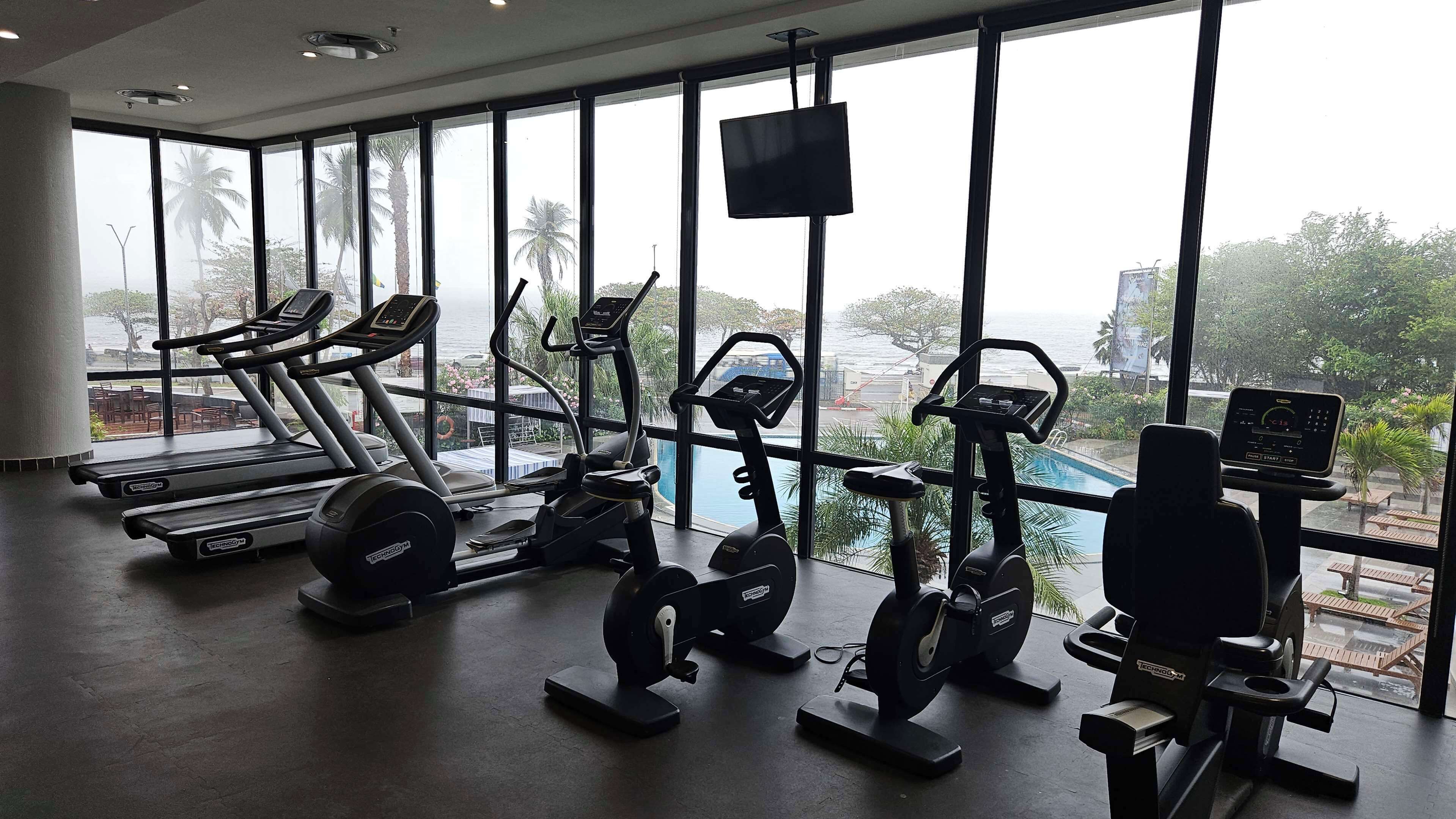 A gym with various exercise machines, large windows, and a view of the outdoors.