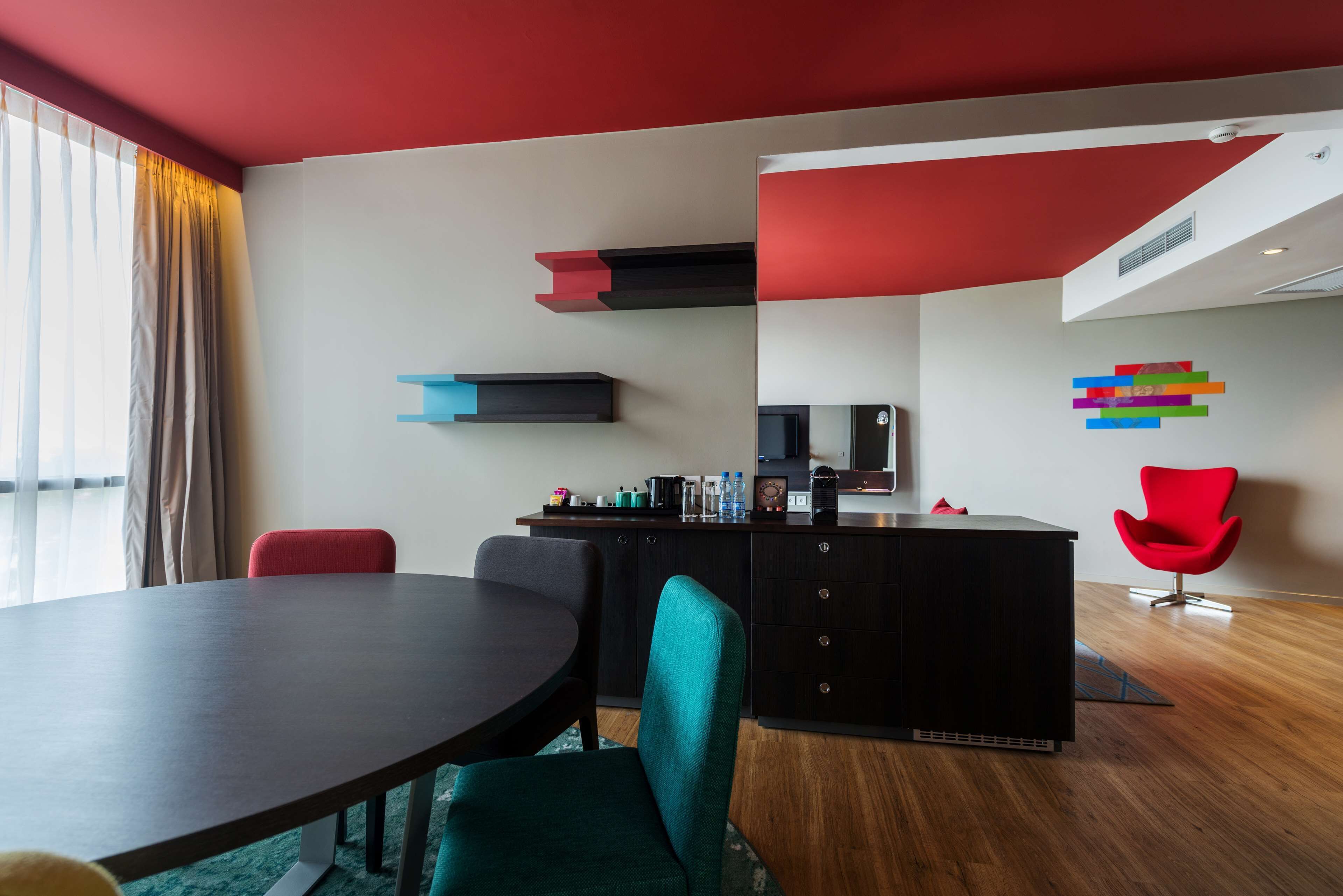A modern hotel room with a dark wooden dining table, teal chairs, a black minibar, and a red accent wall.