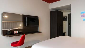 Une chambre d’hôtel moderne avec un grand lit, un fauteuil rouge, un téléviseur à écran plat fixé à un mur sombre, et une petite table avec des livres et une lampe.