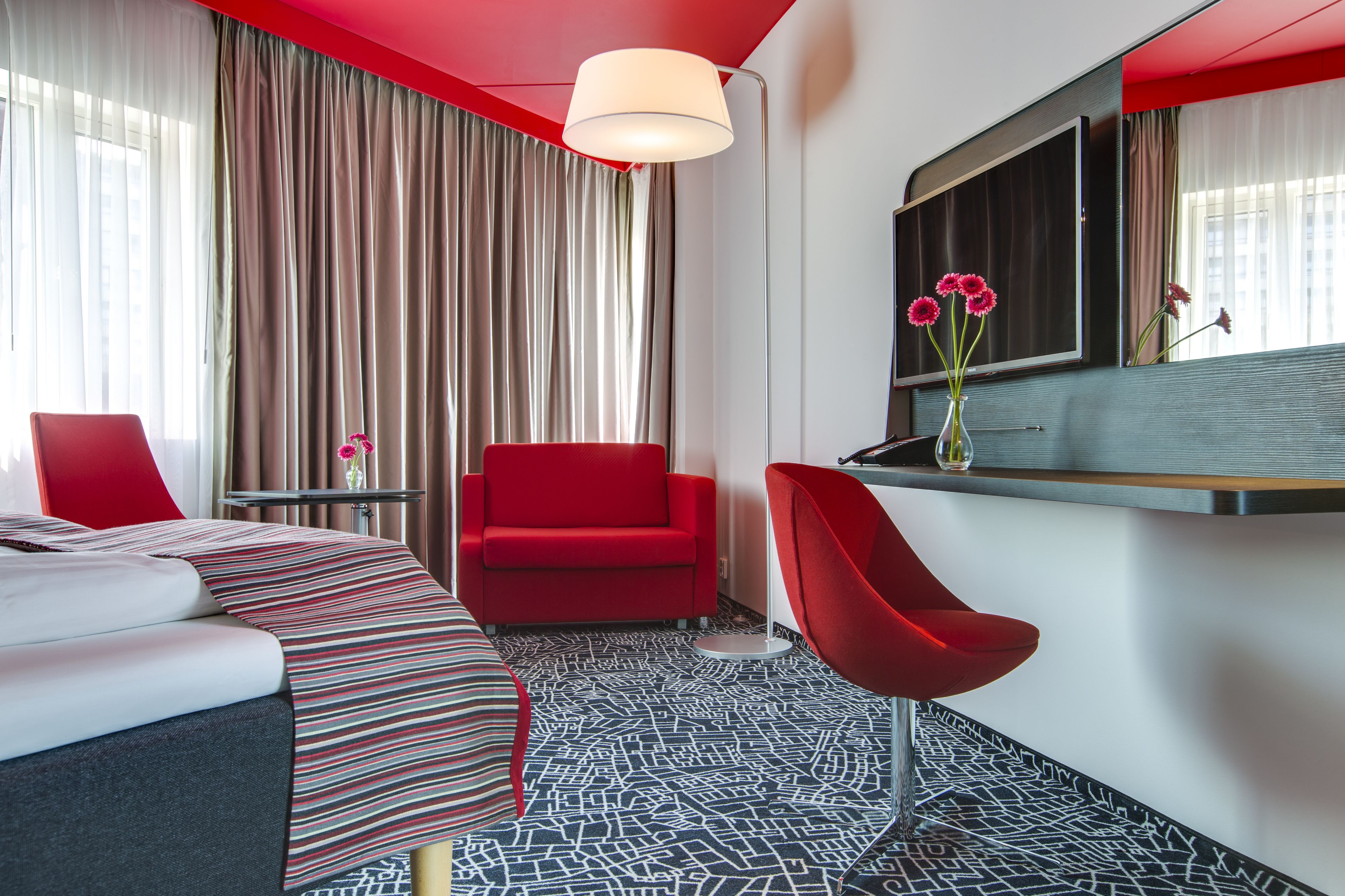 Une chambre d’hôtel moderne avec un lit, un fauteuil rouge, une petite table et un grand miroir.