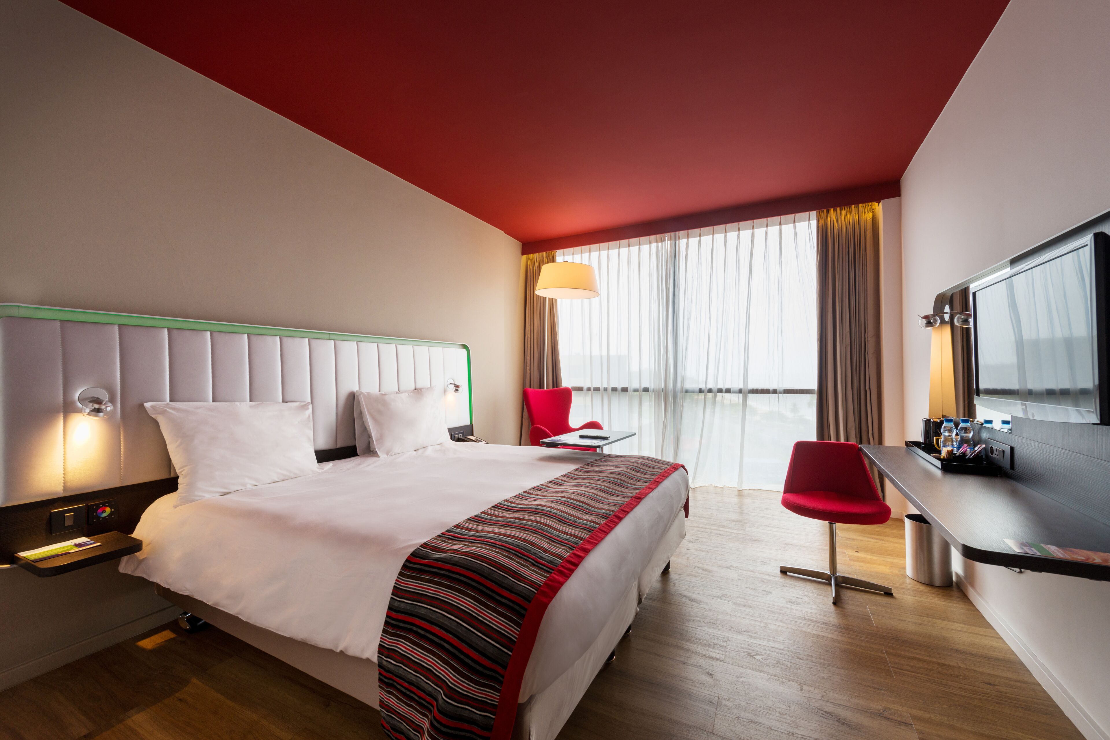 Une chambre d’hôtel avec un lit, un bureau, une chaise rouge et une grande fenêtre avec des rideaux.