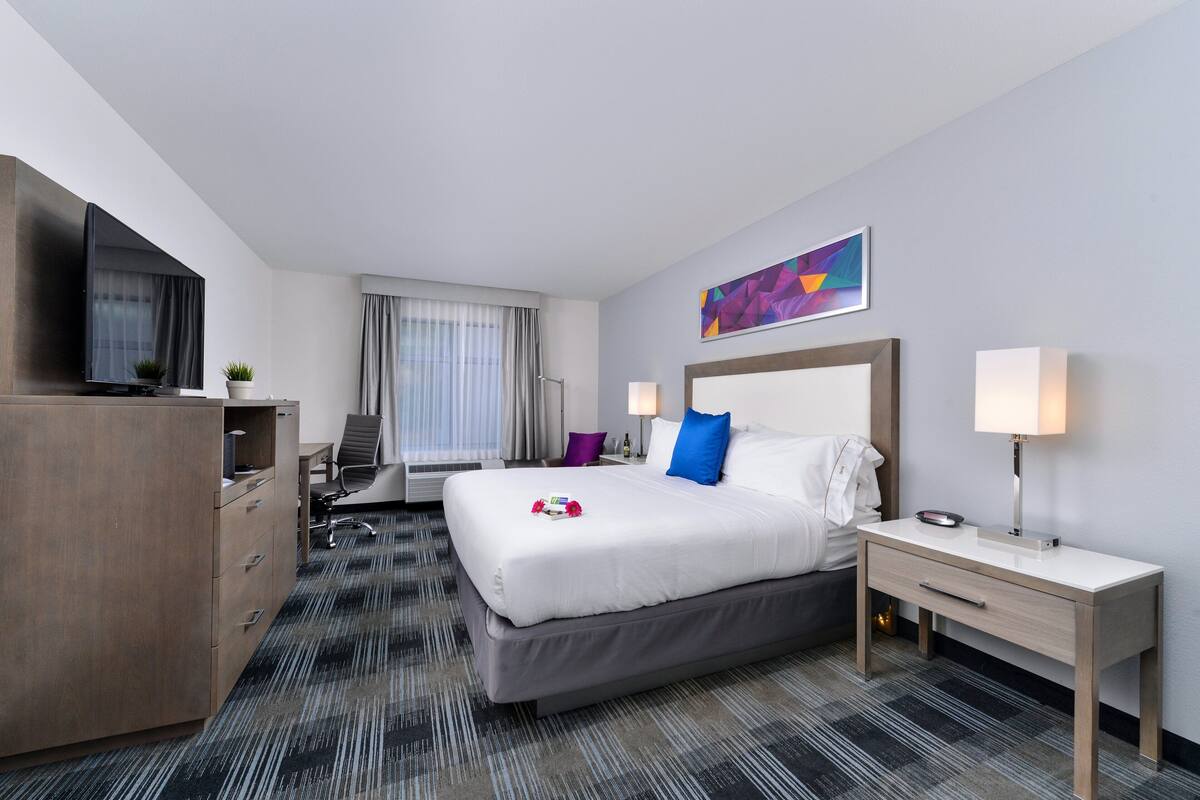 A hotel room with a bed, bedside tables, a desk, a chair, a TV, and a window with curtains.