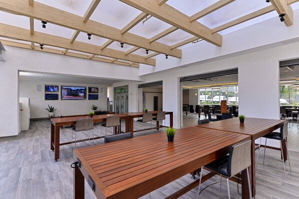 A spacious indoor area with wooden tables and chairs, a high ceiling with wooden beams, and a view of the outside through large windows.