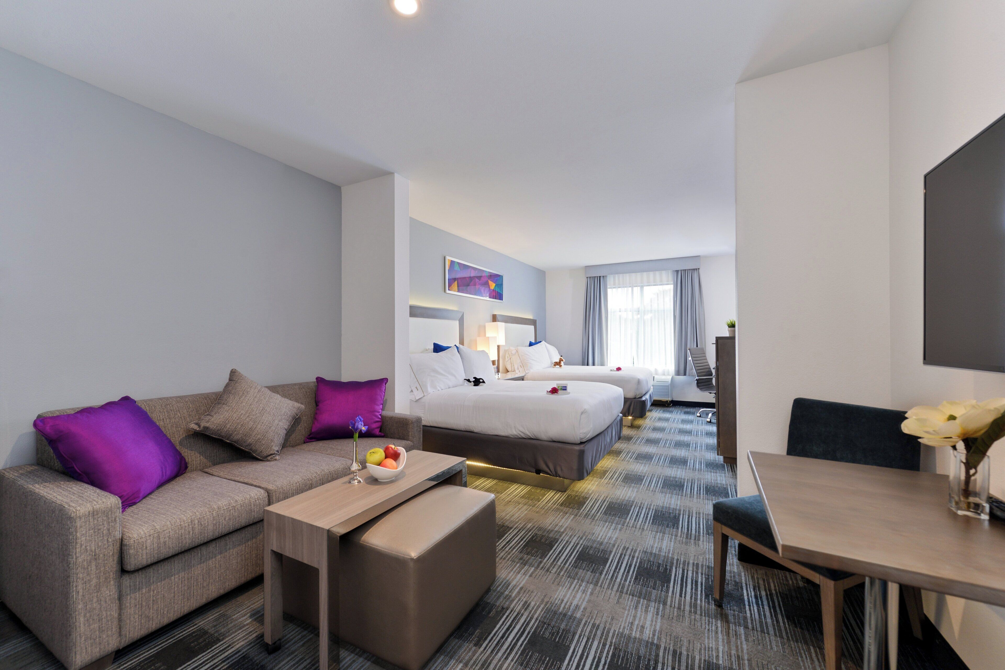 A hotel room with a sofa, a small table, a bed with two pillows, a bedside table, a chair, and a desk with a chair.