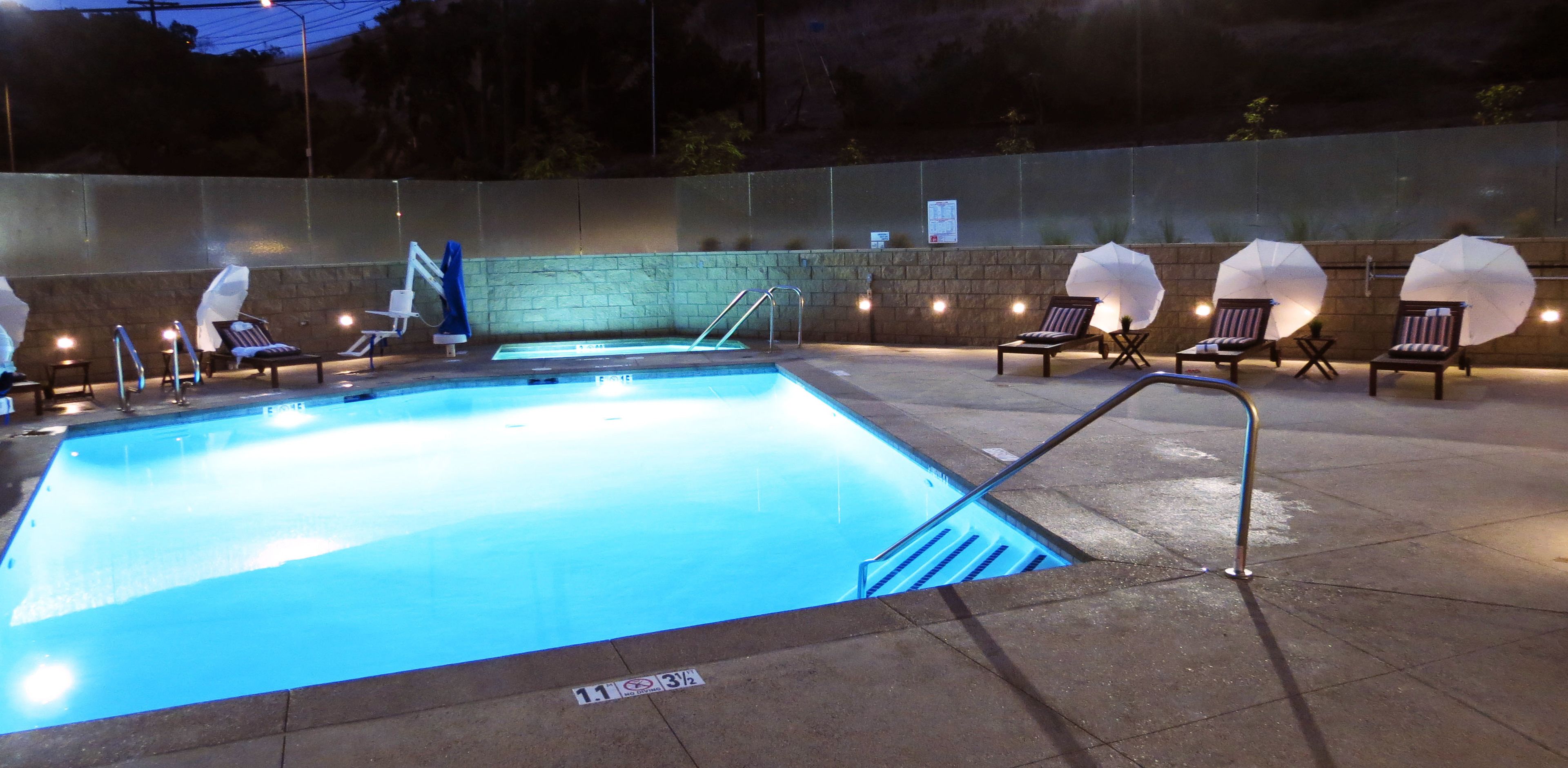 A swimming pool area with lounge chairs and umbrellas, a stone wall, and a pool ladder.
