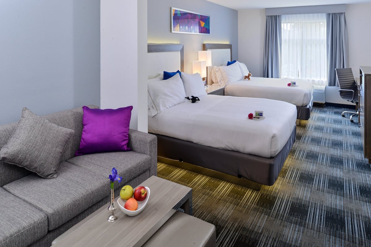 A hotel room with two beds, a sofa, a coffee table with fruit, and a desk with a chair.