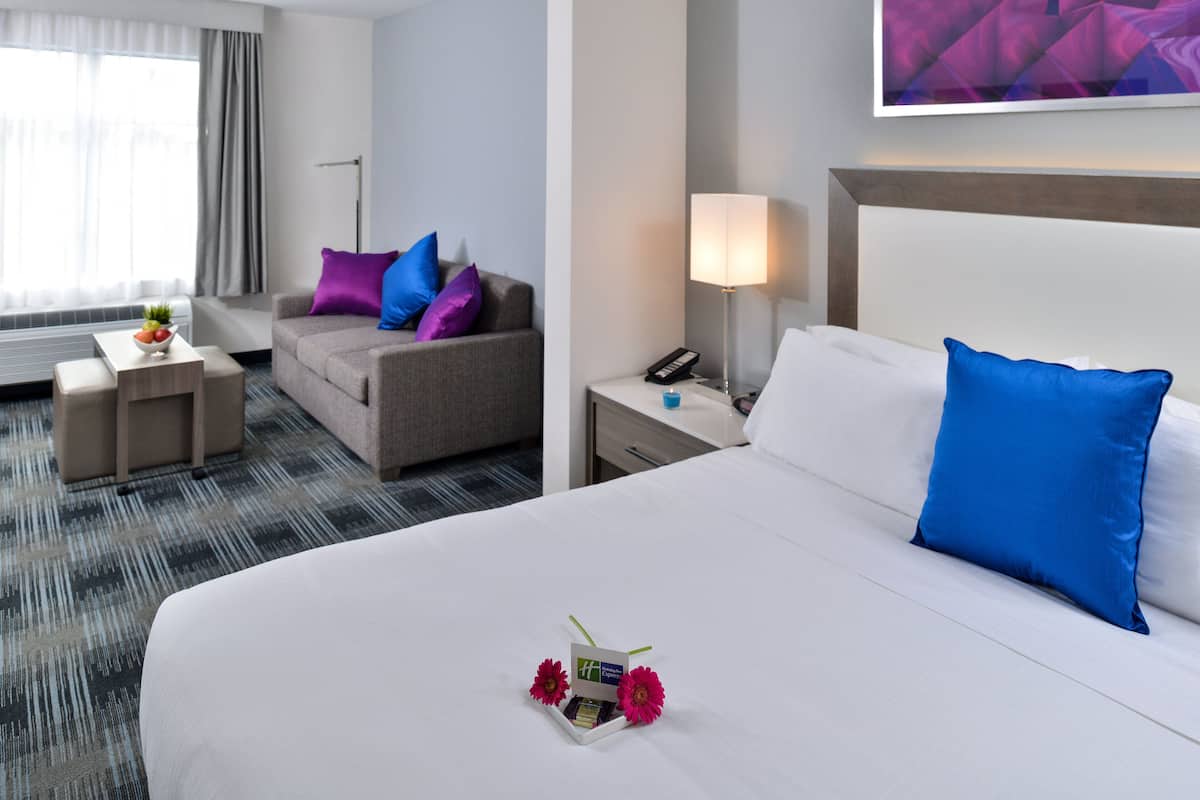 A hotel room with a large bed, a sofa, a small table, a nightstand, and a lamp.