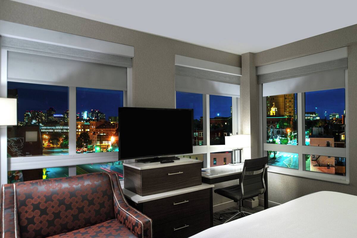 Ein Hotelzimmer mit großem Fenster, einem Flachbildfernseher, einem Schreibtisch, einem Sessel und einer Couch.