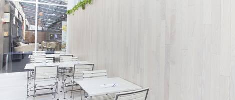 A modern indoor dining area with white tables and metal chairs, a glass ceiling, and a light-colored wooden wall.