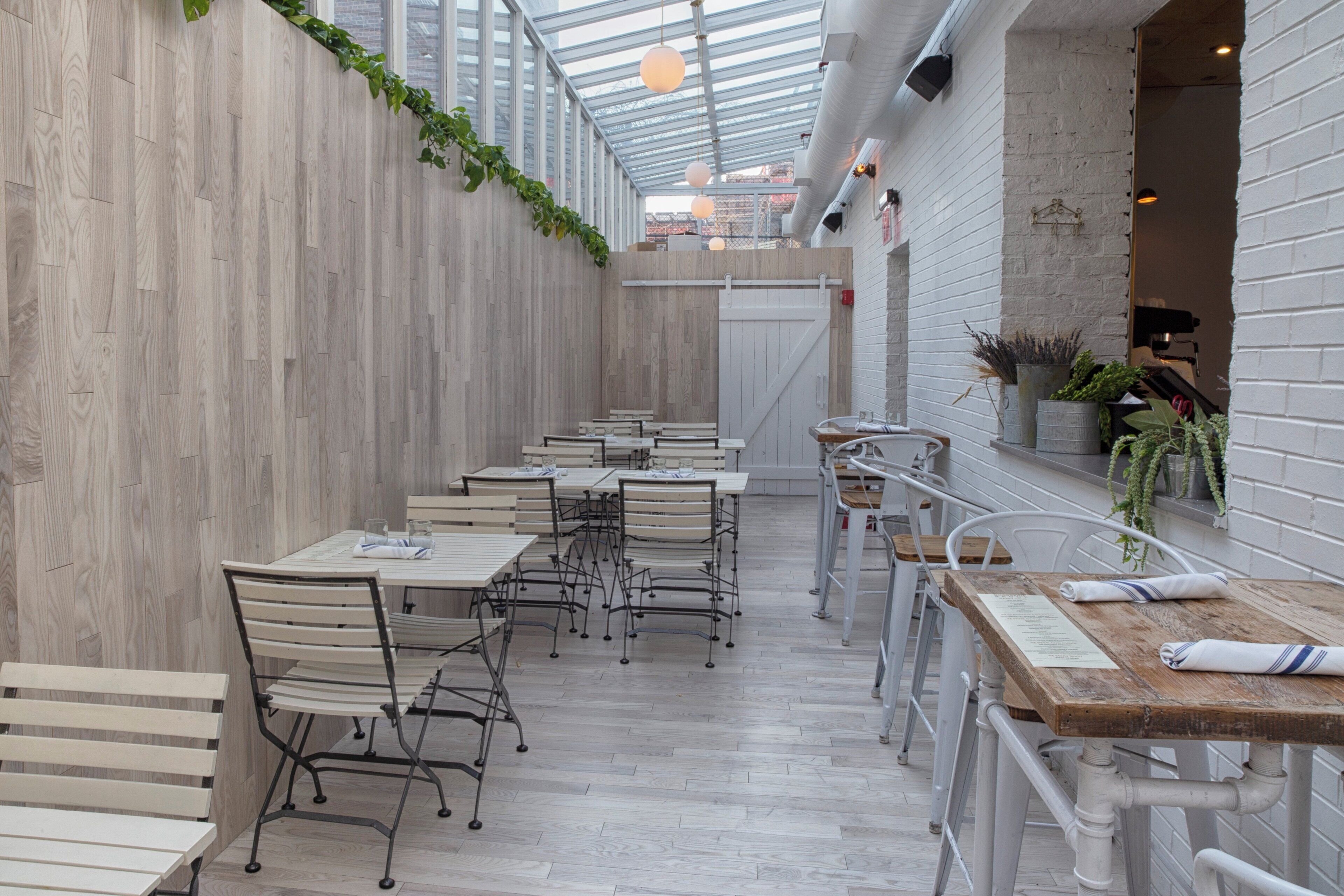 An indoor dining area with wooden flooring, white walls, and a glass ceiling. Tables and chairs are arranged for seating.