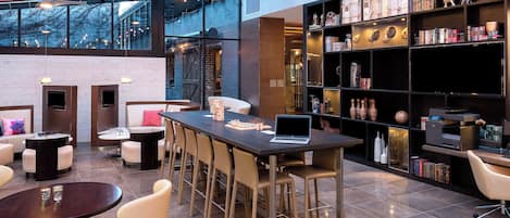 Un espacio lounge moderno con una mesa larga, taburetes de bar, chimenea y estantería con diversos objetos.