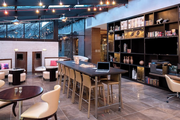 A modern lounge area with a long table, bar stools, a fireplace, and a bookshelf with various items.
