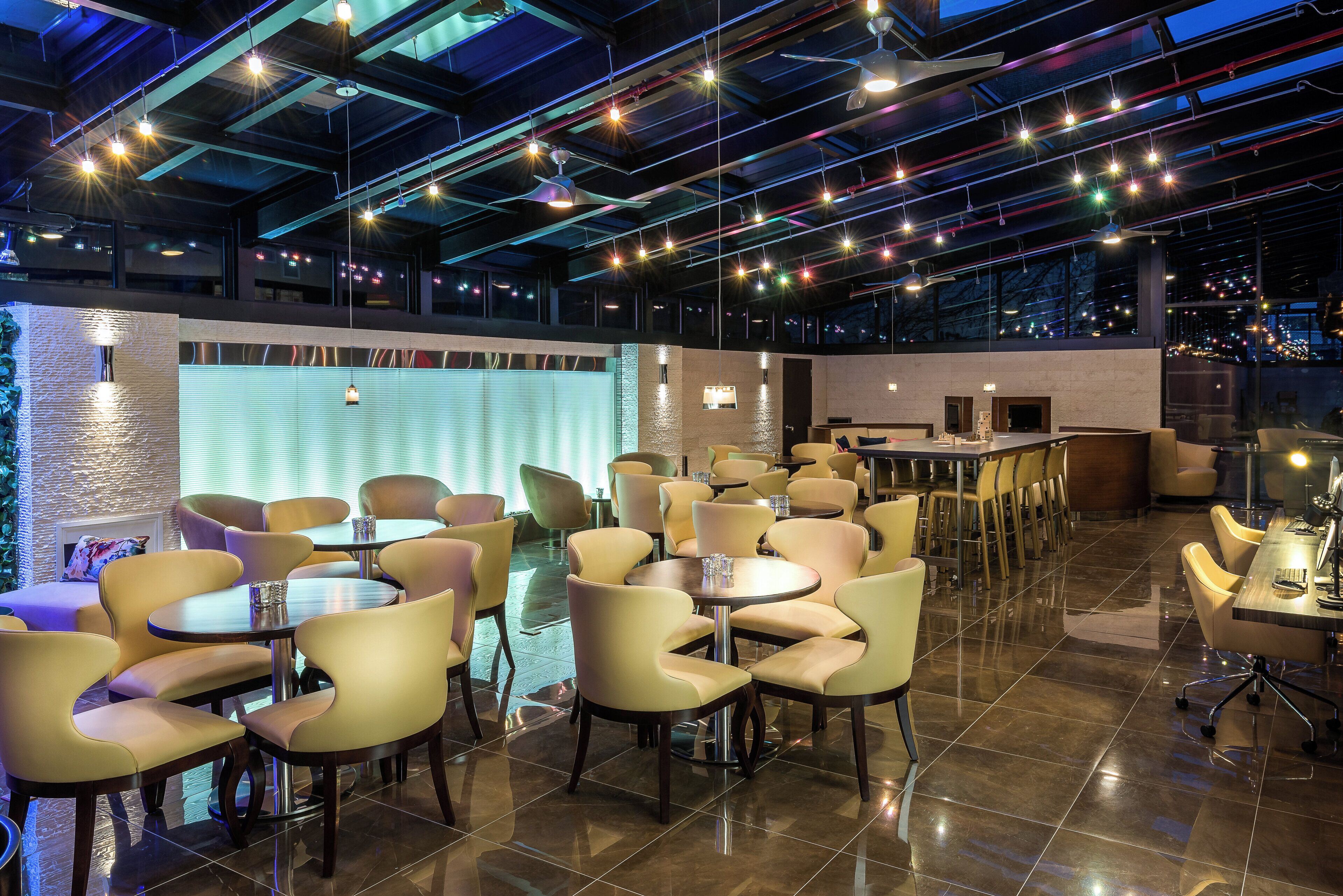 A spacious indoor area with multiple round tables and chairs, a bar area, and a view of the city through large windows.