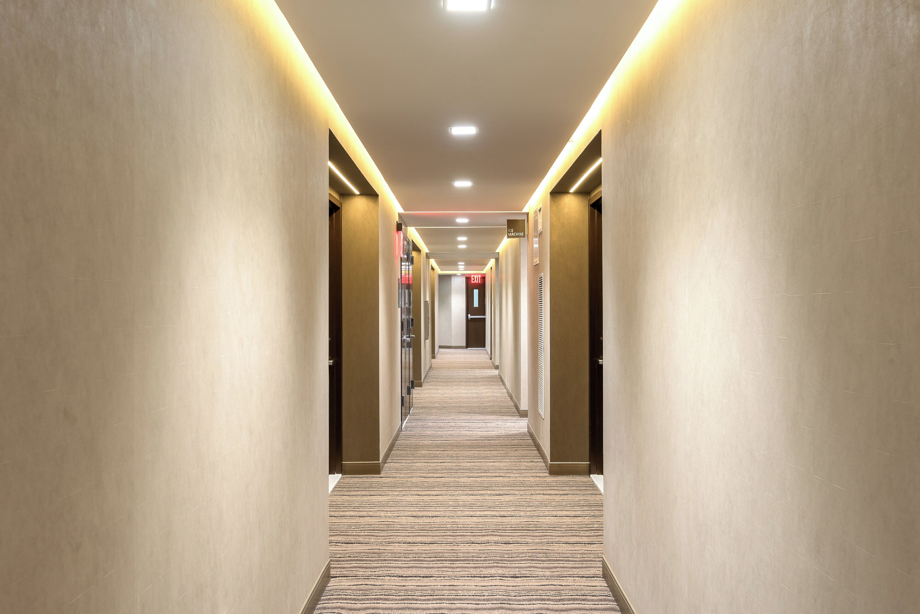 A hotel corridor with a carpeted floor, walls with a light finish, and a ceiling with recessed lighting.