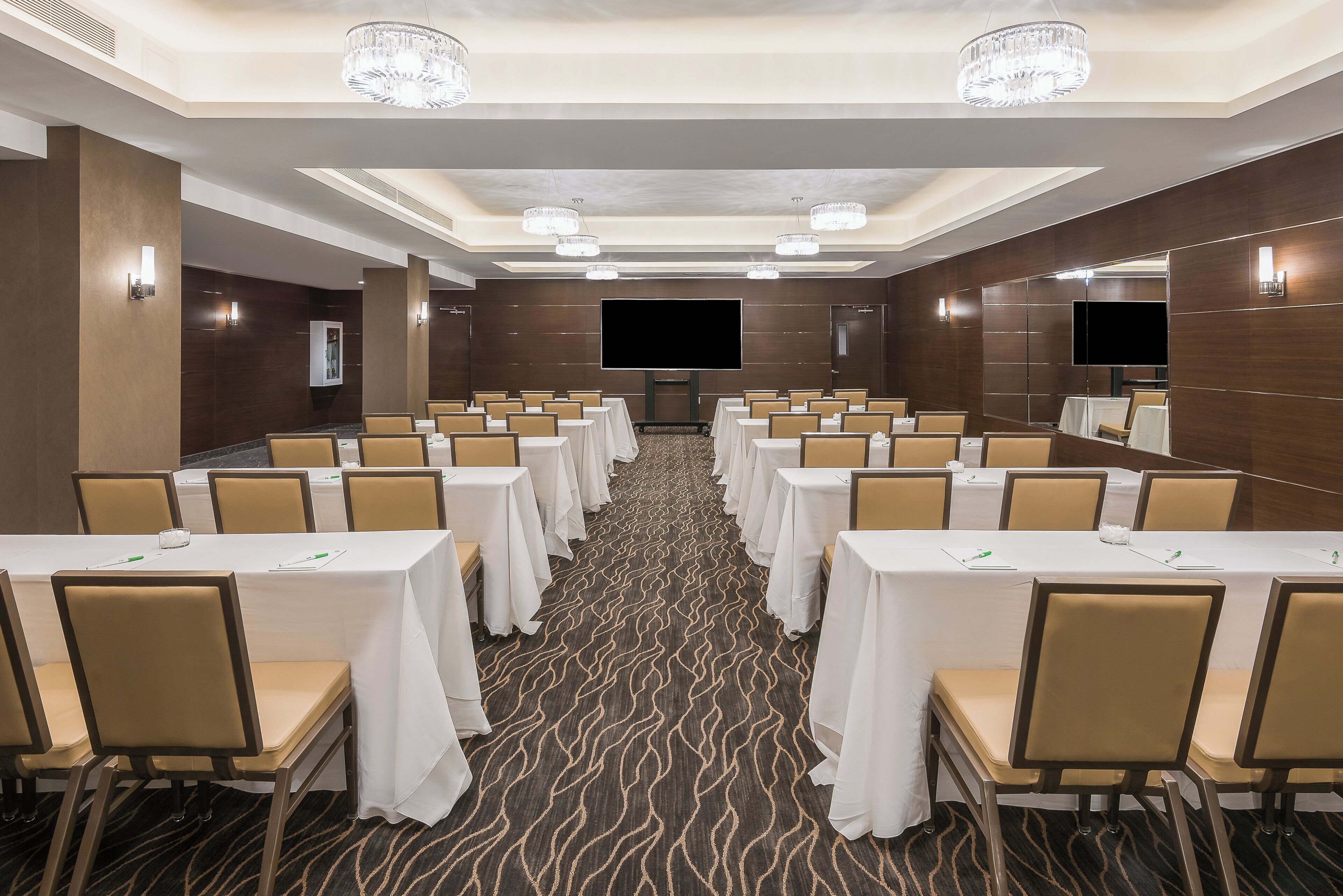A conference room with rows of tables and chairs, a large flat-screen TV, and chandeliers.