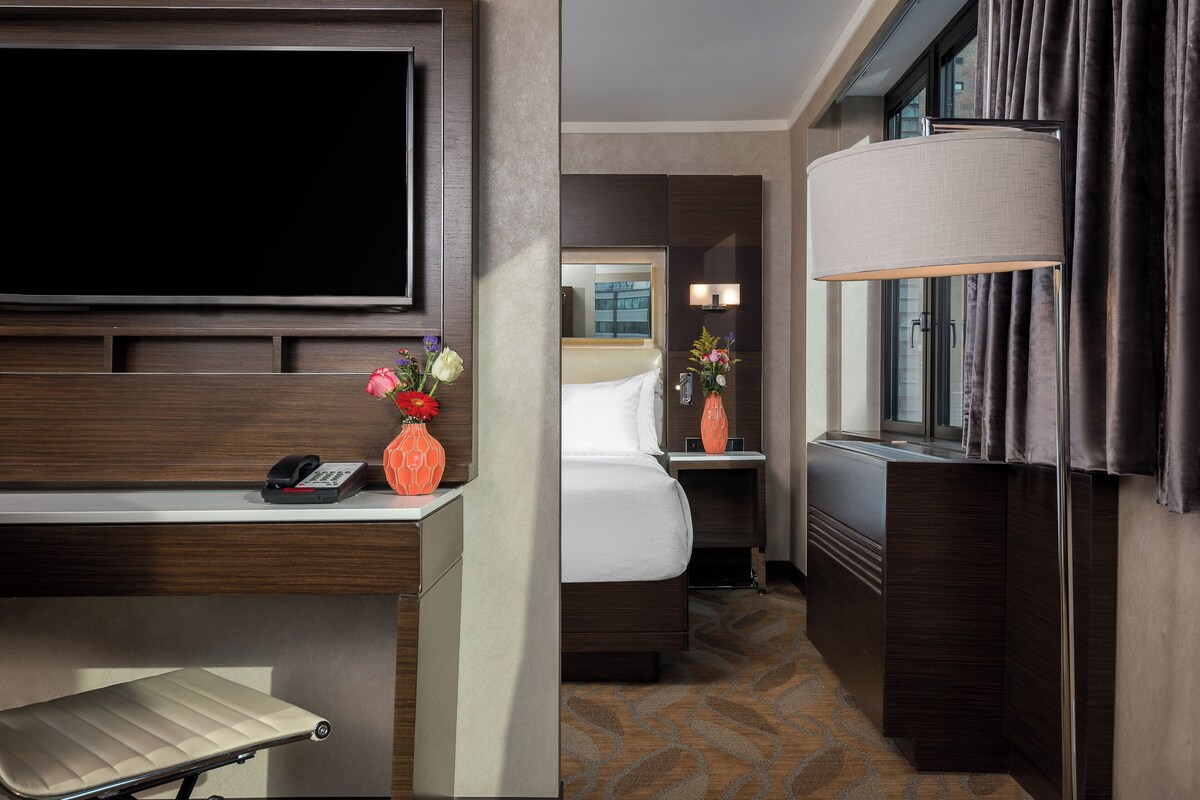 Habitación de hotel con cama, mesita de noche, lámpara y televisión montada en la pared.