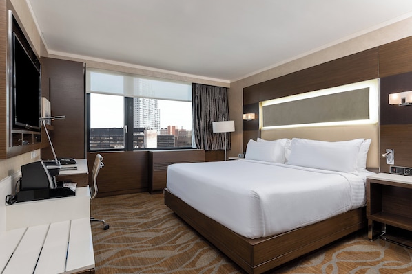 A hotel room with a large bed, a desk with a computer, a TV, and a window with a city view.