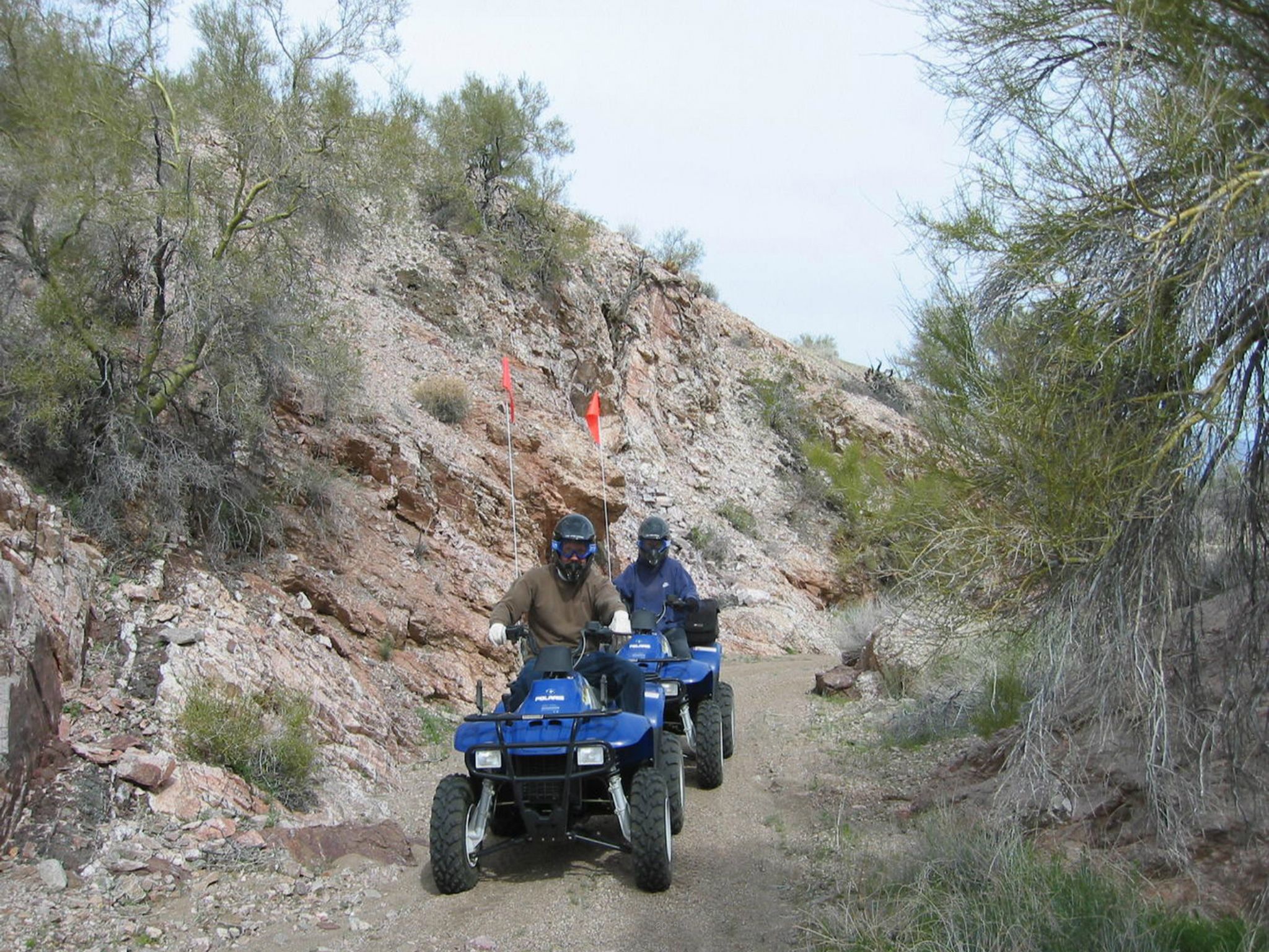 Two people on ATVs on a rocky path with sparse vegetation and a rocky hillside in the background.