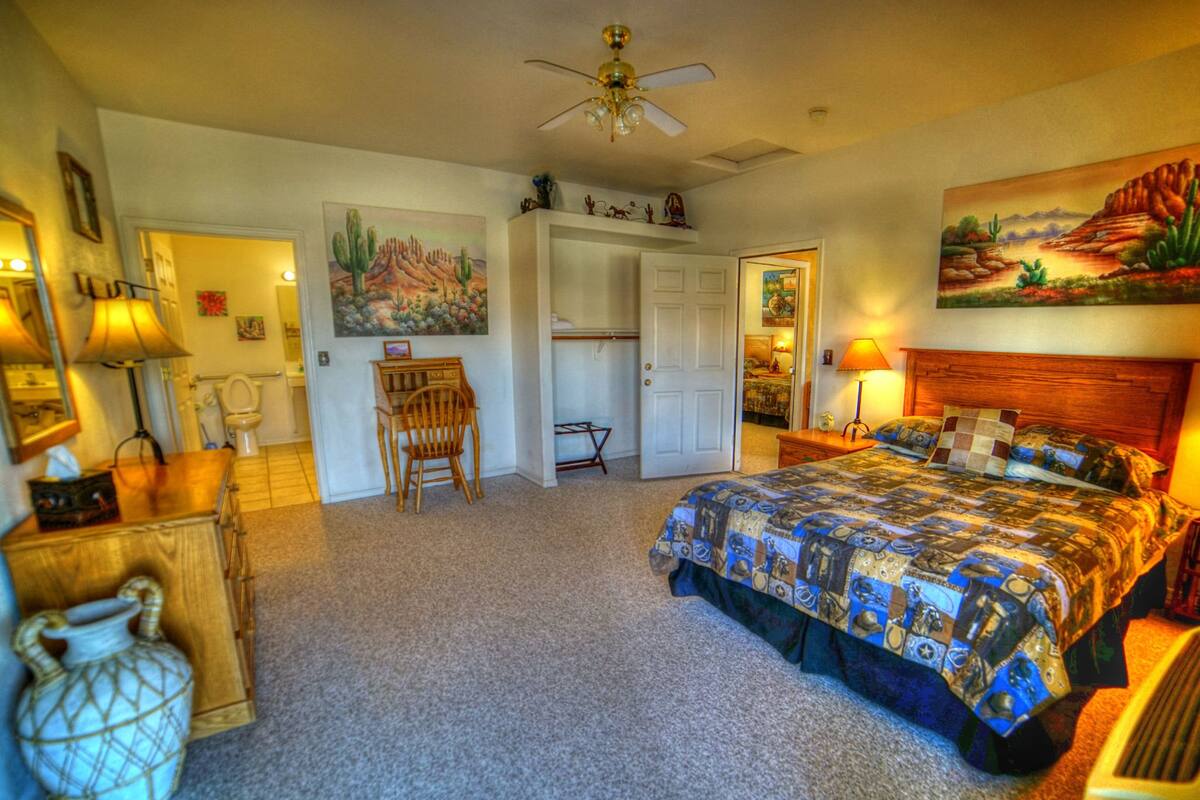 A bedroom with a bed, a desk, a chair, a ceiling fan, and a painting on the wall.