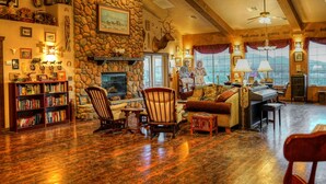 A cozy living room with a stone fireplace, wooden furniture, and a bookshelf filled with books. - Stagecoach Trails Guest Ranch (Yucca)