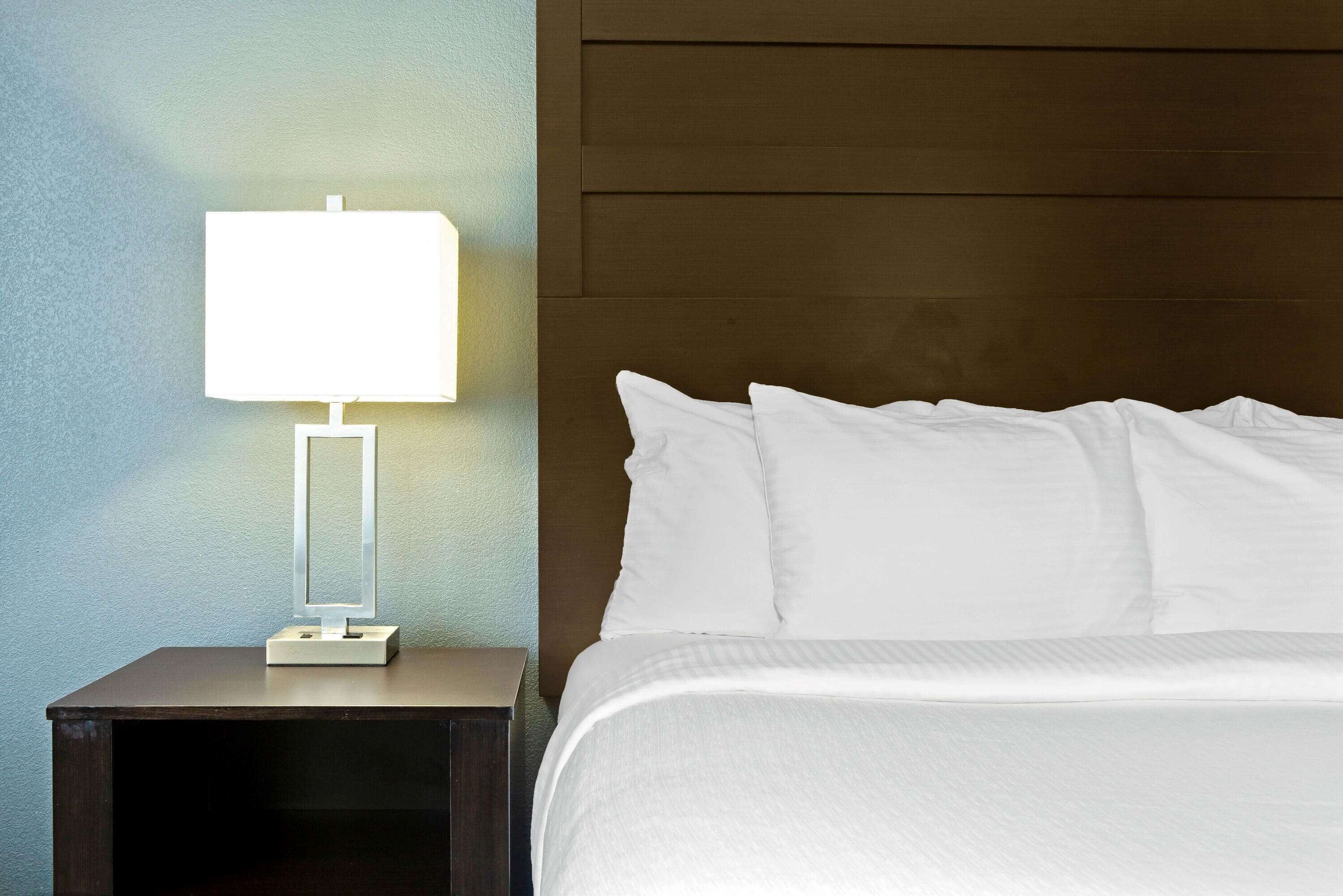 Una cama bien hecha con sábanas blancas, una lámpara de noche sobre una mesita de noche de madera oscura y una pared con textura.