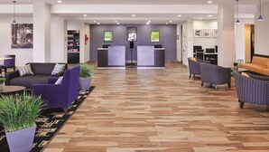Um hall amplo com padrão geométrico em herringbone, balcões de recepção, áreas de estar e um balcão com tela digital.