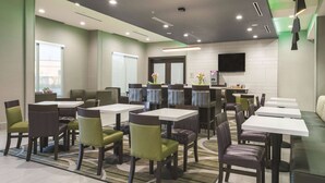 A modern dining area with multiple tables and chairs, a flat-screen TV mounted on the wall, and a large window providing natural light. - La Quinta Inn & Suites by Wyndham Fort Worth West - I-30 (Fort Worth)