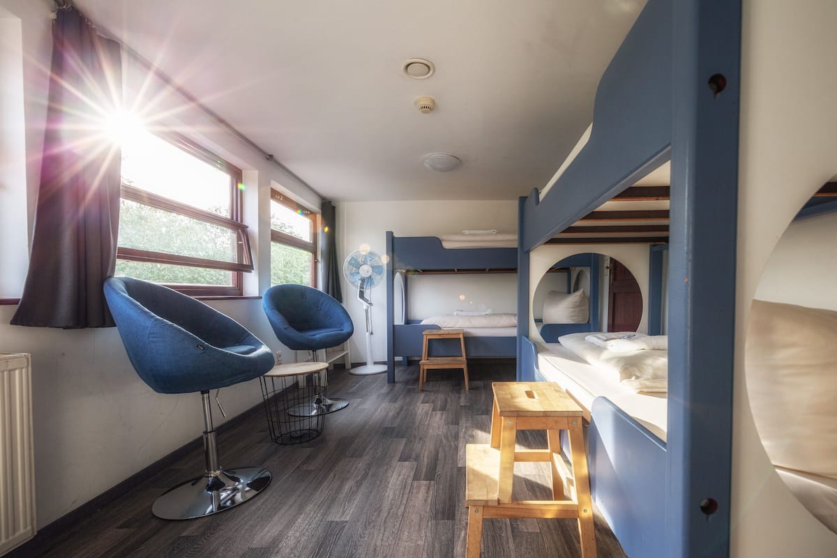 A compact room with a bunk bed, a desk with a chair, a small table, and a fan.