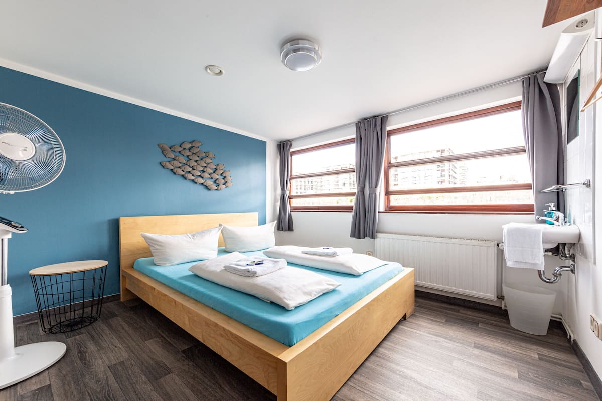 A modern hotel room with a wooden bed, a blue accent wall, a fan, a round table, and a view of the city through large windows.